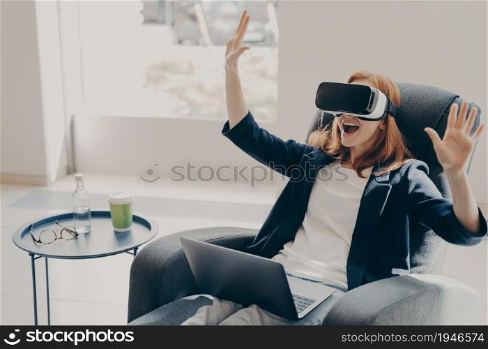 Young excited redhead woman testing VR glasses or goggles while sitting on armchair at home, working remotely. Impressed female with opened mouth using VR technology for the first time, gesturing. Young excited redhead woman testing VR glasses or goggles while sitting on armchair at home