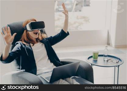 Young excited redhead woman testing VR glasses or goggles while sitting on armchair at home, working remotely. Impressed female with opened mouth using VR technology for the first time, gesturing. Young excited redhead woman testing VR glasses or goggles while sitting on armchair at home