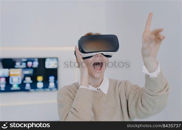 Young excited woman in VR headset in office. Girl in VR goggles is pointing with finger touching virtual buttons. Businesswoman working on project in cyberspace. Futuristic business concept.. Young excited woman in VR headset pointing with finger touching virtual buttons.