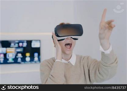 Young excited woman in VR headset in office. Girl in VR goggles is pointing with finger touching virtual buttons. Businesswoman working on project in cyberspace. Futuristic business concept.. Young excited woman in VR headset pointing with finger touching virtual buttons.