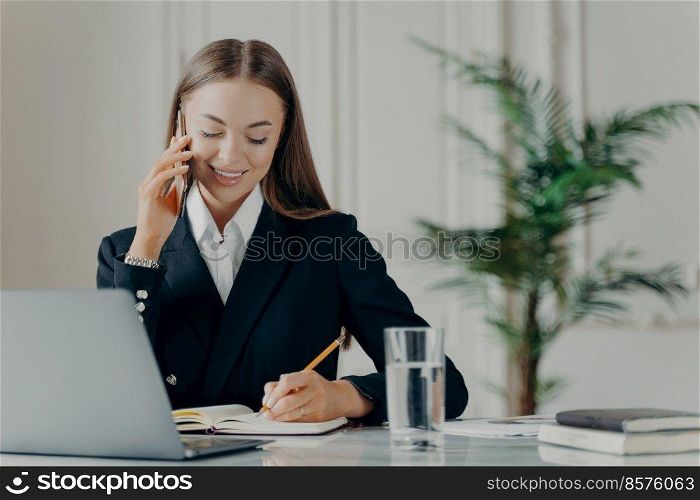 Young experienced business consultant in elegant black suit talking on phone, writing notes with pencil in notepad while communicating with client, female manager working on laptop at her workplace. Positive young businesswoman talking on phone and writing notes