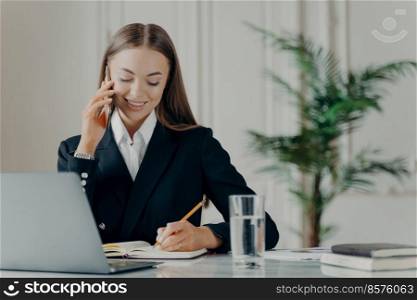 Young experienced business consultant in elegant black suit talking on phone, writing notes with pencil in notepad while communicating with client, female manager working on laptop at her workplace. Positive young businesswoman talking on phone and writing notes