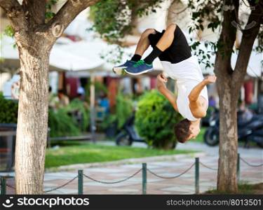 Young extreme athlete doing front flip between the trees. City street with outdoor cafe in background. Young sportsman doing front flip in the street