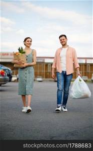 Young family couple with cardboard bags on supermarket car parking. Happy customers carrying purchases from the shopping center, vehicles on background. Young couple with bags on supermarket parking