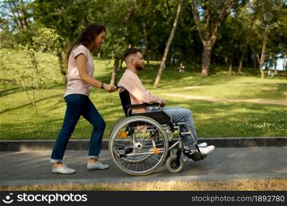 Young family couple with wheelchair walking in the park. Paralyzed people and disability, care for a disabled man. Husband and wife overcome difficulties together, warm relationships. Young couple with wheelchair walking in the park