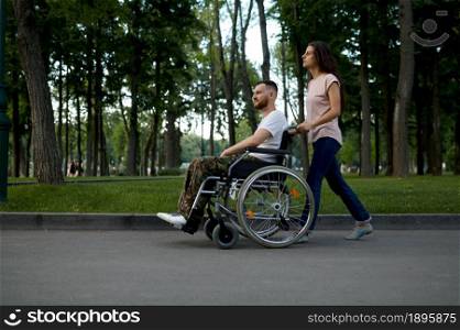Young family couple with wheelchair walking in the park. Paralyzed veteran and disability, care for a disabled man. Husband and wife overcome difficulties together, warm relationships. Young couple with wheelchair walking in the park