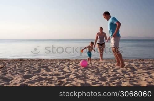 Young family frolicking together playing football soccer on the beach at sunset with the little boy running with his mother and father