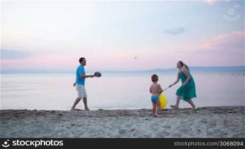Young family having fun relaxing at the seaside in the evening with the parents playing with bats and a ball tennis while their little son carries a yellow beach ball with smile across the sand