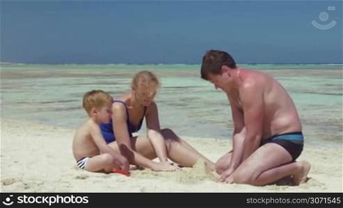 Young family is resting by the sea. Parents are helping their son in building a sand castle.