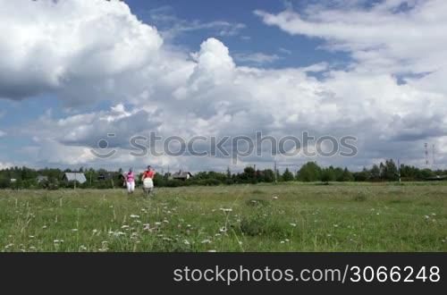 Young family of three in shorts and T-shirts walking with their buggy outdoors across the field on a sunny day. Wide shot. Shot with polarizer.
