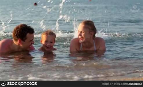 Young family splashing in the sea with attractive parents flanking their small son lying in the shallow surf in the summer sun
