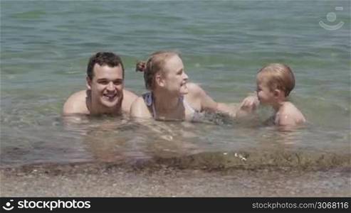Young family splashing in the sea with their small son lying in the shallow surf in the summer sun