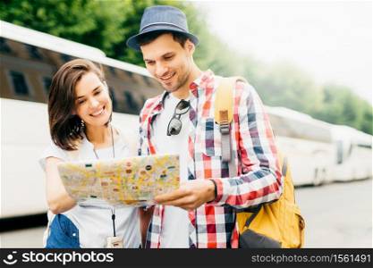 Young family tourists looking attentively in map while searching for museum in big city. Stylish man in hat and checkered shirt holding rucksack, standing near his wife, looking for new destination