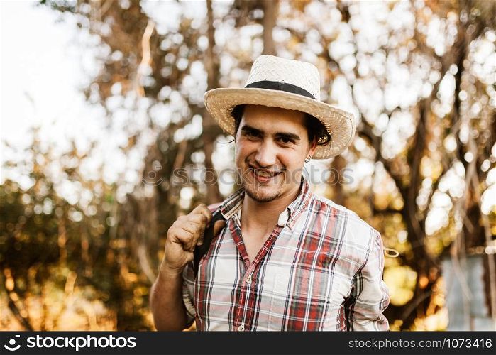 Young farmer gets ready to work in field with manual pump tank wearing an old hat and plaid shirt