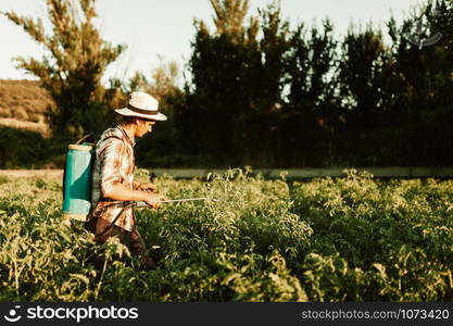 Young farmer spraying organic fertilizer with manual pump tank wearing an old hat and plaid shirt