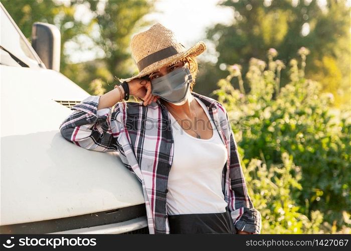 Young farmer woman with straw hat and surgical mask leaning against the van and holding her hat