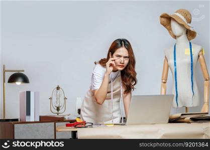 Young fashion designer or tailor wearing eyeglasses with working with laptop computer standing with feels displeasure gesture, full of tailoring tools with mannequin and equipment on desk in studio