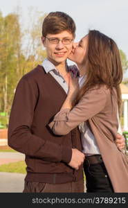 Young fashion elegant stylish couple in love posing in a European city park. Cute girl kisses a hipster man.