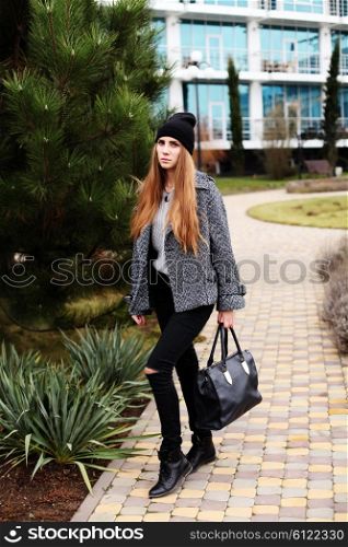 Young fashion sensual woman in black hat, warm gray coat with leather bag posing outdoor