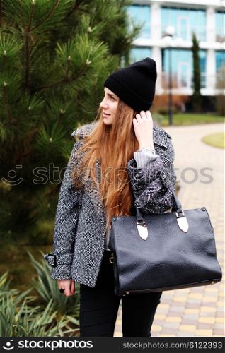 Young fashion sensual woman in black hat, warm gray coat with leather bag posing outdoor