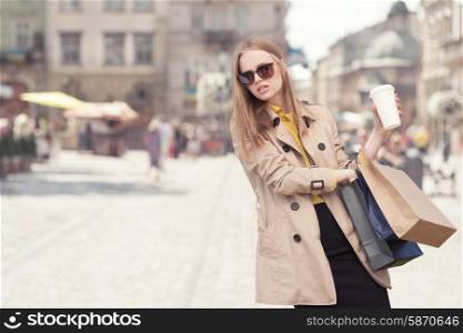 Young fashionable woman carrying heavy shopping bags after shopping, walking with a coffee-to-go in her hands against urban city background.
