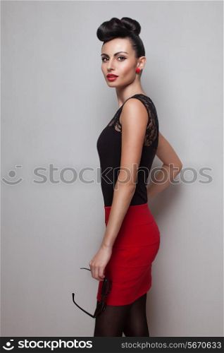 Young fashionable woman holding round sunglasses posing in red skirt. Red lips. Updo, twisted high bun. top knot