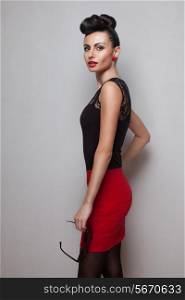 Young fashionable woman holding round sunglasses posing in red skirt. Red lips. Updo, twisted high bun. top knot