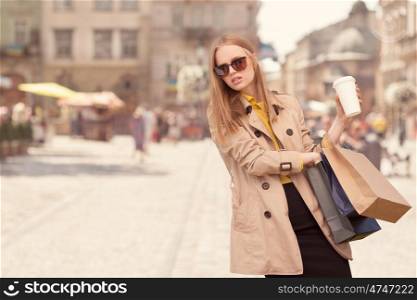 Young fashionable woman taking a coffee break after shopping, walking with a coffee-to-go in her hands against urban city background.