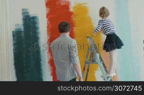 Young father and his little daughter are painting the wall in different colors. Girl is touching dad&acute;s face with brush.