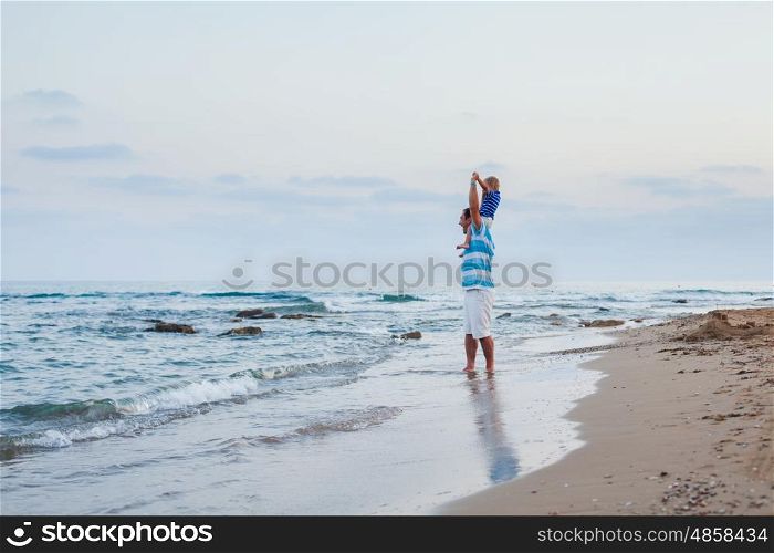 Young father and his little son spending time outdoors in summer time walking along sand beach. Son sits on his father's shoulders and having fun