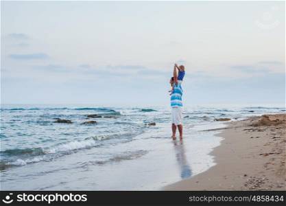 Young father and his little son spending time outdoors in summer time walking along sand beach. Son sits on his father's shoulders and having fun