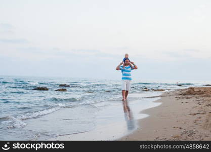 Young father and his little son spending time outdoors in summer time walking along sand beach. Son sits on his father's shoulders and having fun