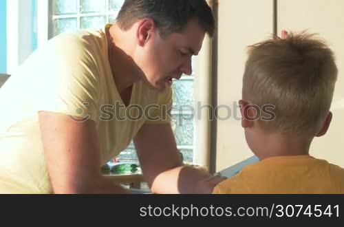 Young father and little son are sitting on the open veranda in sunny day, man is reading a book aloud to the boy.