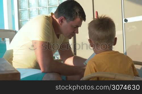 Young father and little son spending leisure time on the balcony. Father reading story to child and showing pictures in the book