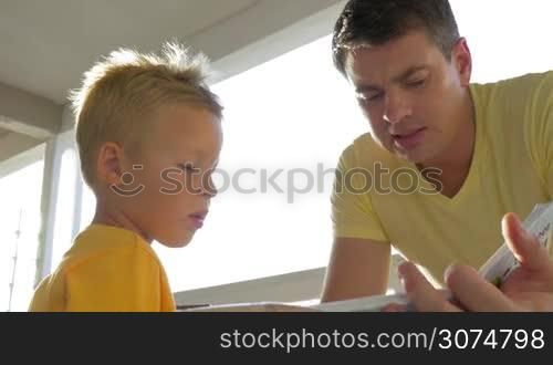 Young father and little son spending leisure time reading a book on the balcony on sunny day. Child listening to dad very attentively