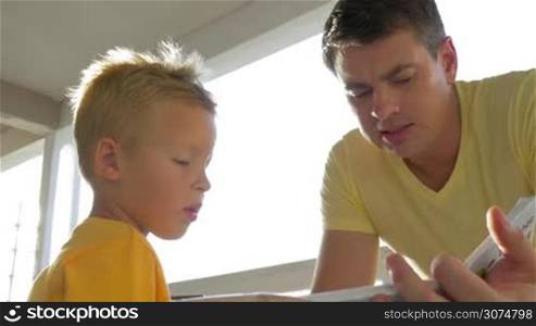 Young father and little son spending leisure time reading a book on the balcony on sunny day. Child listening to dad very attentively