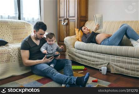 Young father looking at the tablet with his little son while the pregnant mother reads a magazine. Father looking tablet with little son while the pregnant mother reads a magazine