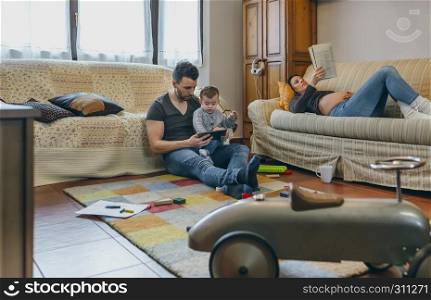 Young father looking at the tablet with his little son while the pregnant mother reads a magazine. Father looking tablet with little son while the pregnant mother reads a magazine