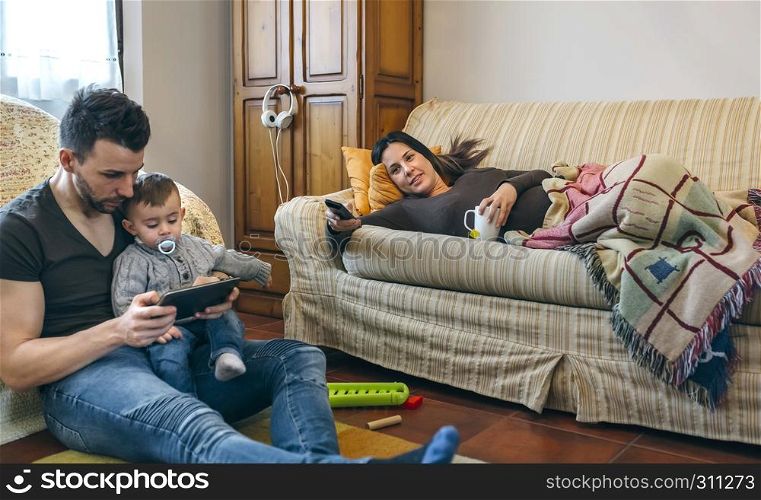 Young father looking at the tablet with his little son while the pregnant mother watches television. Father looking tablet with little son while the pregnant mother watches television
