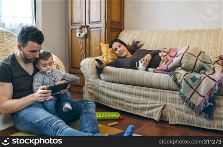 Young father looking at the tablet with his little son while the pregnant mother watches television. Father looking tablet with little son while the pregnant mother watches television