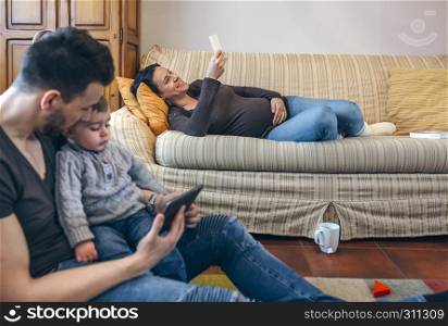 Young father looking at the tablet with his little son while the pregnant mother looks the mobile. Focus on pregnant woman in background. Father looking tablet with little son while the pregnant mother looks the mobile