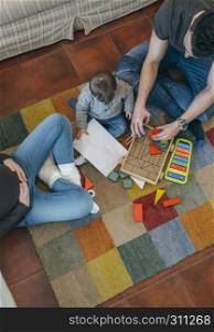 Young father playing with their toddler a wooden game building in the living room while the mother is looking at them. Father playing with toddler while the mother is looking at them
