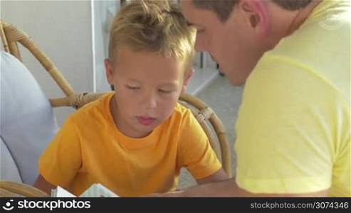 Young father reading a book to the little son. Child sitting in the arm-chair on the balcony and interacting with dad