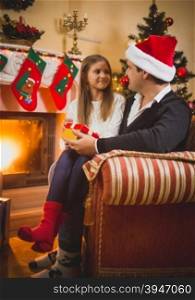 Young father sitting at fireplace giving Christmas present to daughter