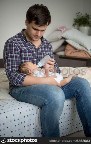 Young father sitting on bed and feeding his newborn son