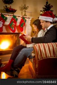 Young father sitting with daughter at fireplace and telling story on Christmas eve