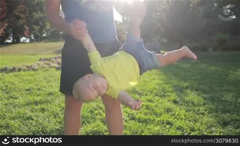 Young father swinging his giggling infant baby boy upside down in summer in glow of beautiful sunset. Playful dad and cute toddler son having fun and playing outdoors over colorful landscape background. Slow motion. Steadicam stabilized shot.
