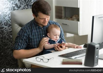 Young father working at computer playing with his baby son