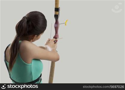 Young female archer aiming arrow against gray background