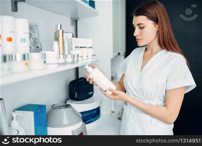 Young female beautician with bottle of cream in hands, cosmetology cabinet. Doctor in white uniform in spa salon, beauty medicine. Female beautician with bottle of cream in hands